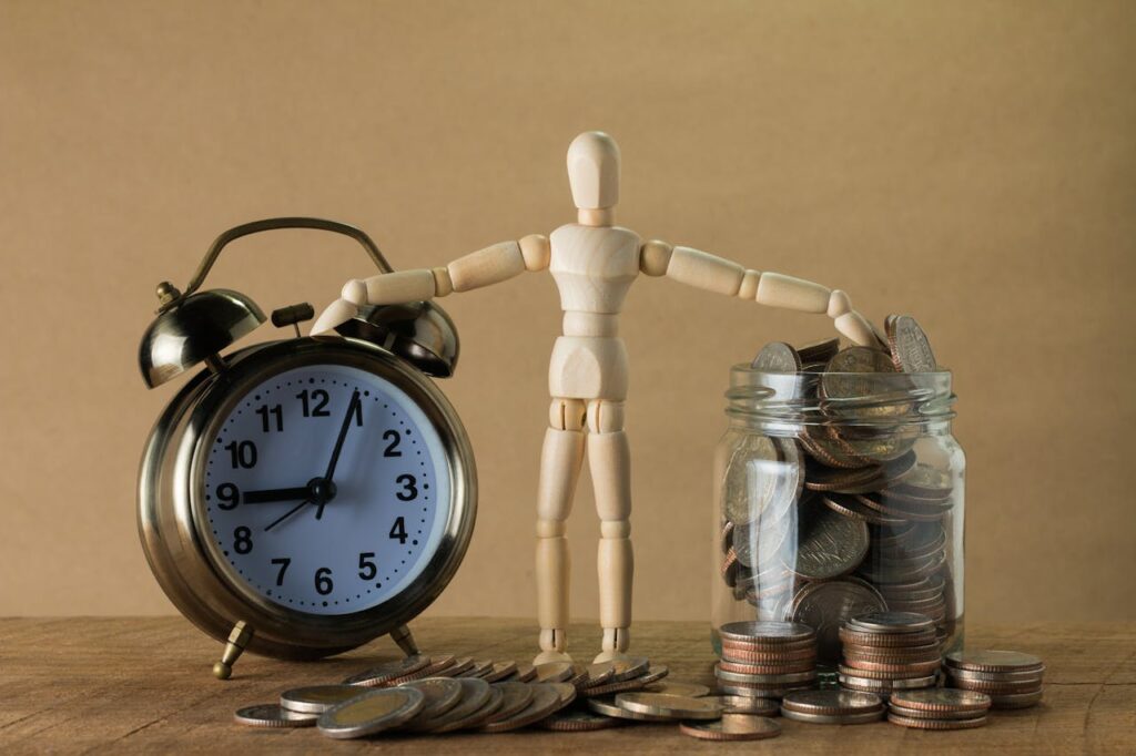 Wooden mannequin holding an alarm clock and a jar full of coins, symbolizing time and money.